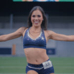 "Cheerleader in navy blue uniform performing with silver pom-poms on a football field, smiling with arms outstretched, in a stadium environment."