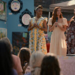 "Four women stand on a stage at an outdoor art fair, three with microphones addressing an audience. Colorful paintings and art are displayed on a blue wall behind them. The crowd is seated in the foreground."